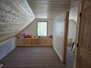 An attic room with angled wood-paneled walls, carpeted floor, built-in wooden storage, and a doorway leading to a hallway.