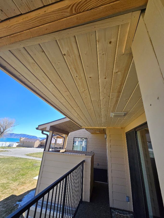A view from a balcony showing a light-wood slat ceiling, metal railing, and a neighboring building under a blue sky.