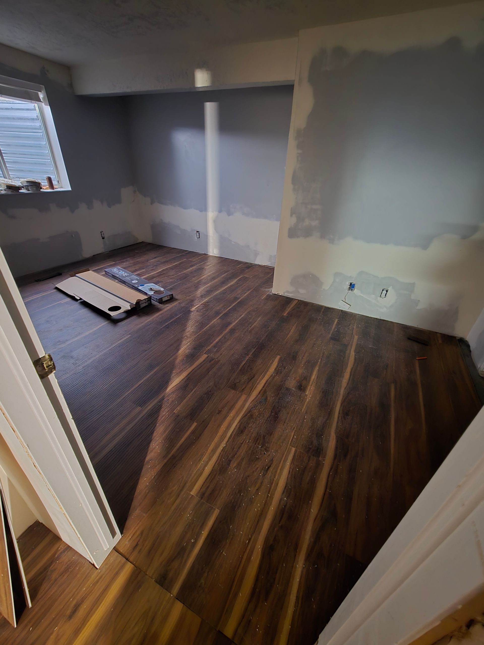 A room under renovation with newly installed dark wood laminate flooring, partially painted walls, and a window.