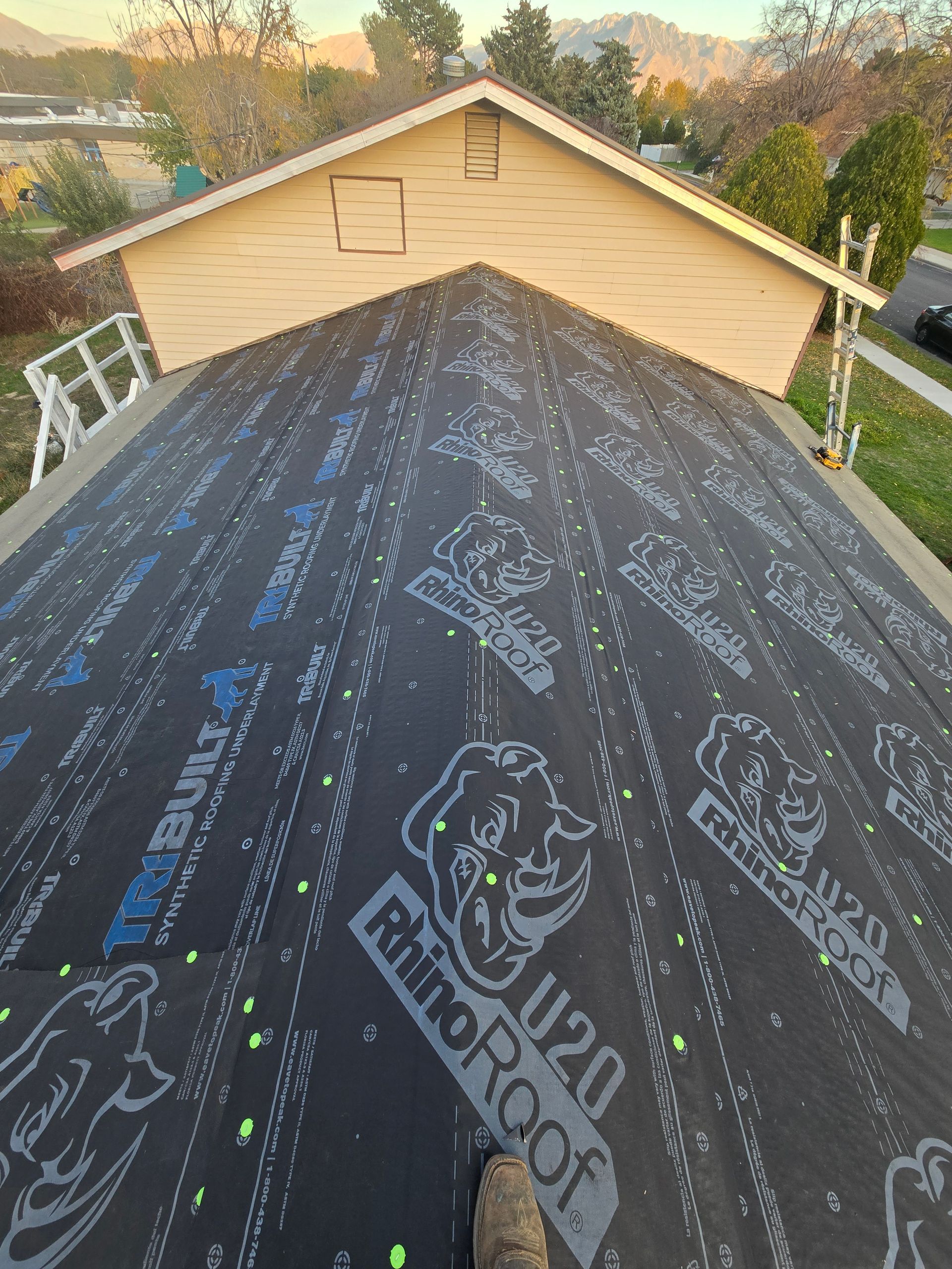 A view from a roof covered in black RhinoRoof underlayment, looking up toward a beige house gable under a clear sky.