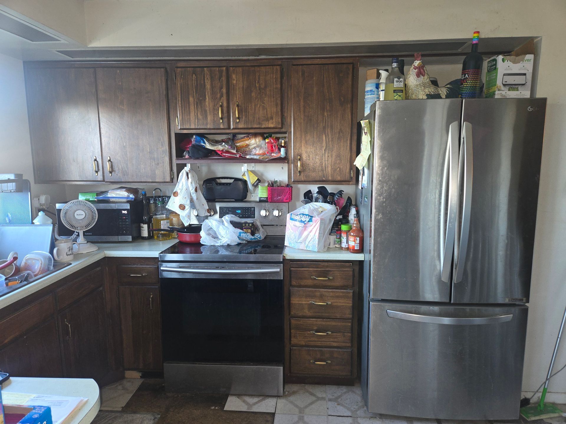 A kitchen featuring dark wood cabinets, a stainless steel oven and refrigerator, and cluttered countertops.