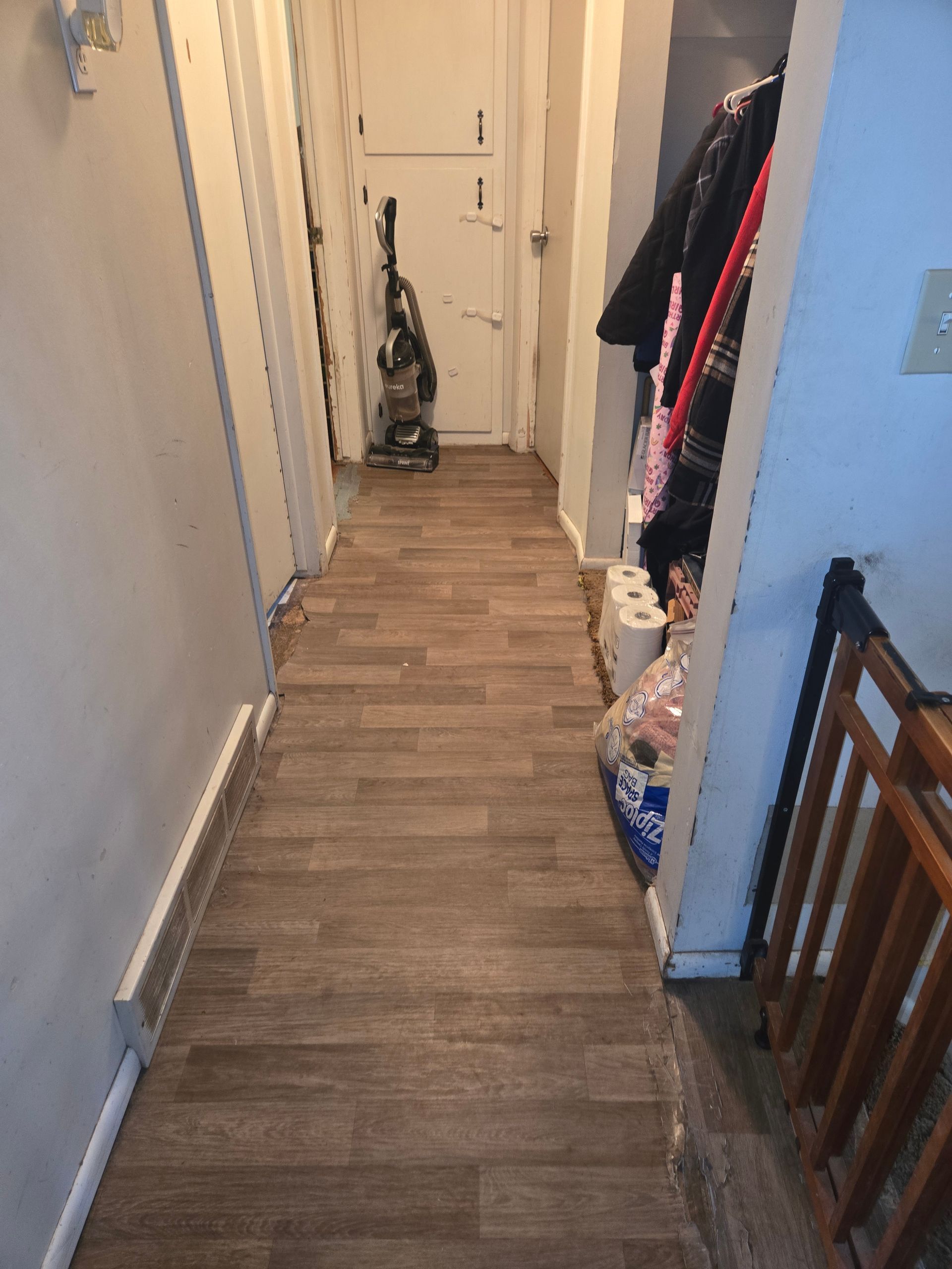 A narrow hallway with wood-look vinyl flooring, featuring a vacuum near a white door and coats hanging in a side alcove.
