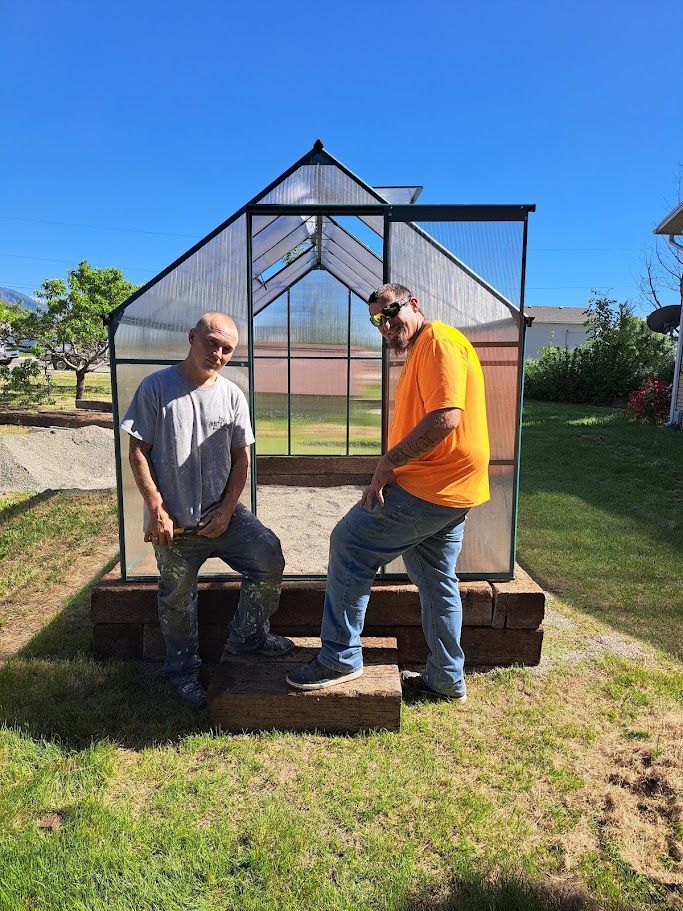 Two people standing in front of a newly assembled greenhouse in a grassy backyard on a sunny day.