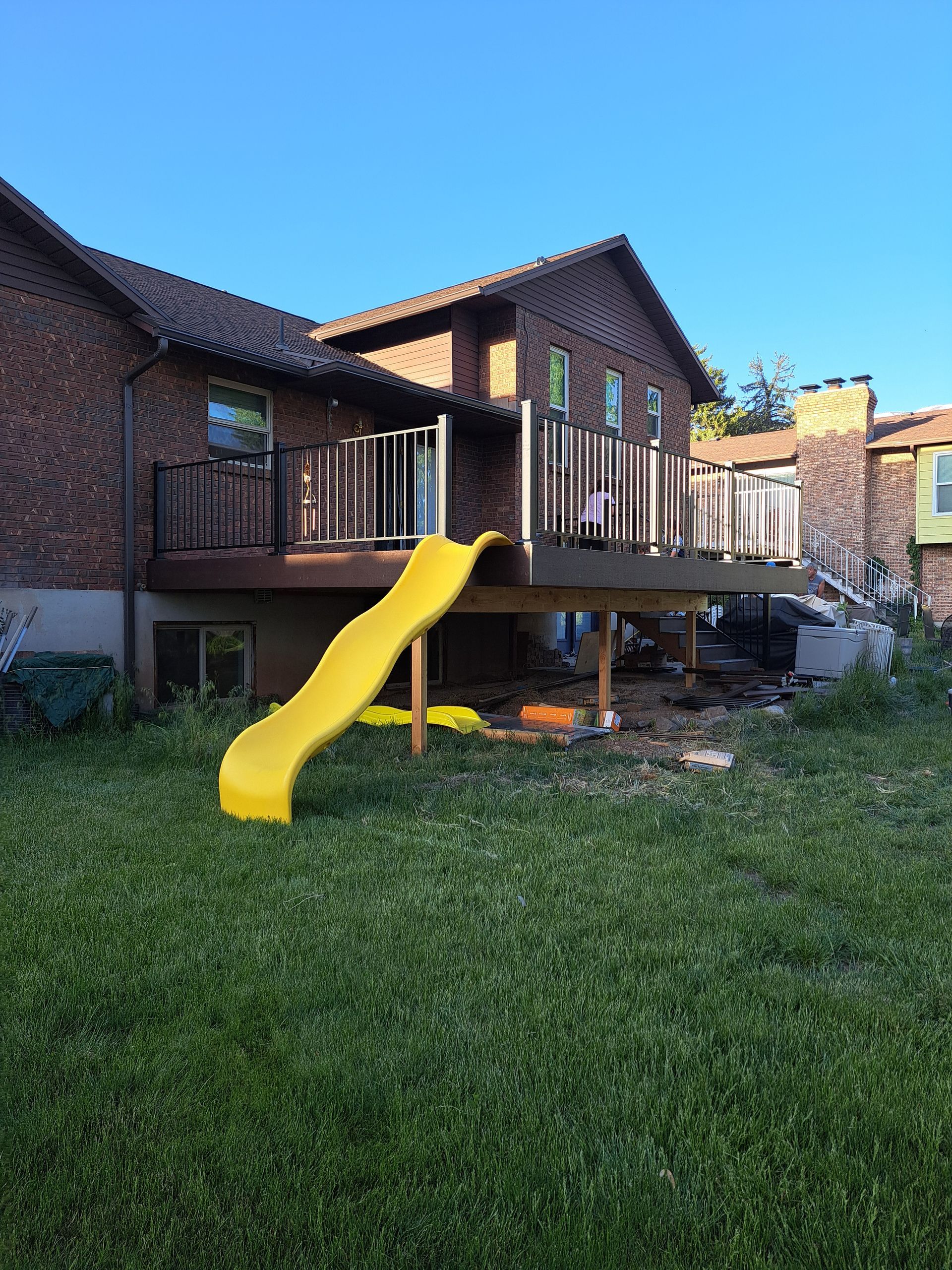 A brick house with an elevated wooden deck and a bright yellow slide leading down to a green backyard.