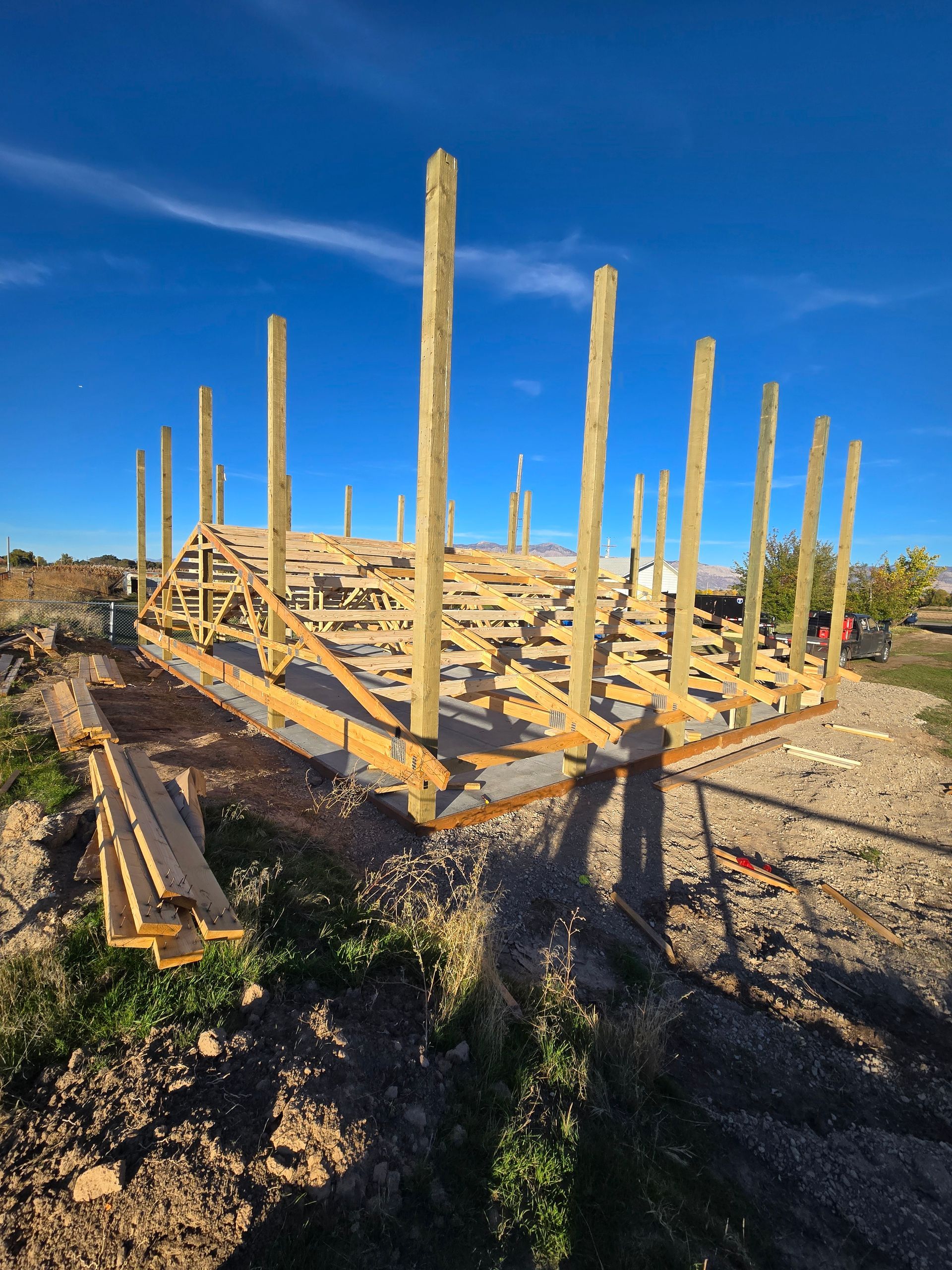A wooden pole barn frame under construction on a dirt lot against a clear blue sky.