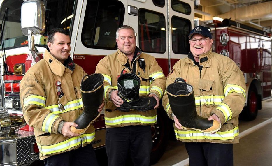 Three firefighters holding up boots in front of a fire engine.