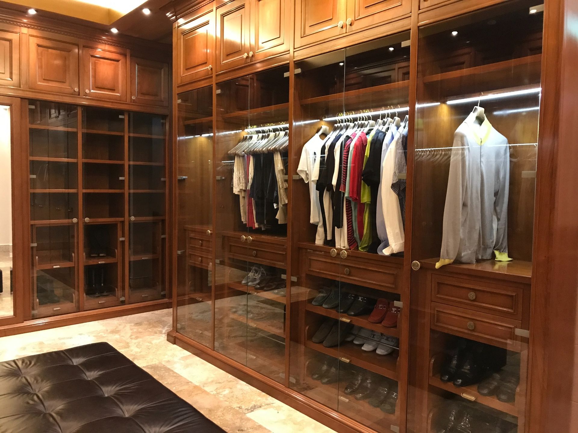 Walk-in closet with brown wood cabinets, glass doors, hanging clothes, and a black bench.