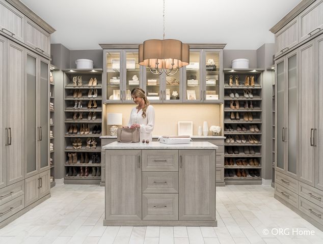 Woman in walk-in closet with island, selecting items. Gray cabinets, shoe shelves, and warm lighting.