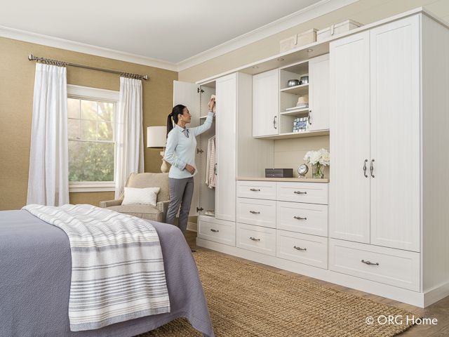 Woman reaching into white closet in bedroom. Wardrobe, drawers, bed, window.