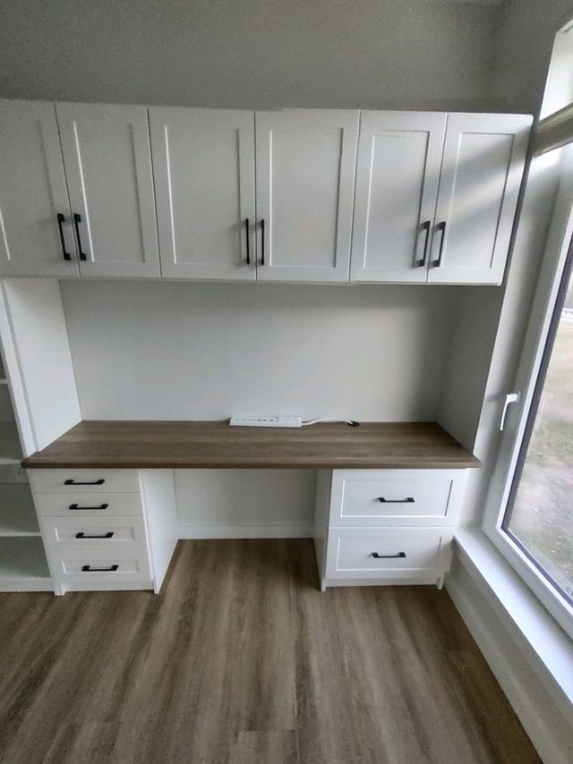 Built-in white desk with overhead cabinets, brown countertop, drawers, and a window.