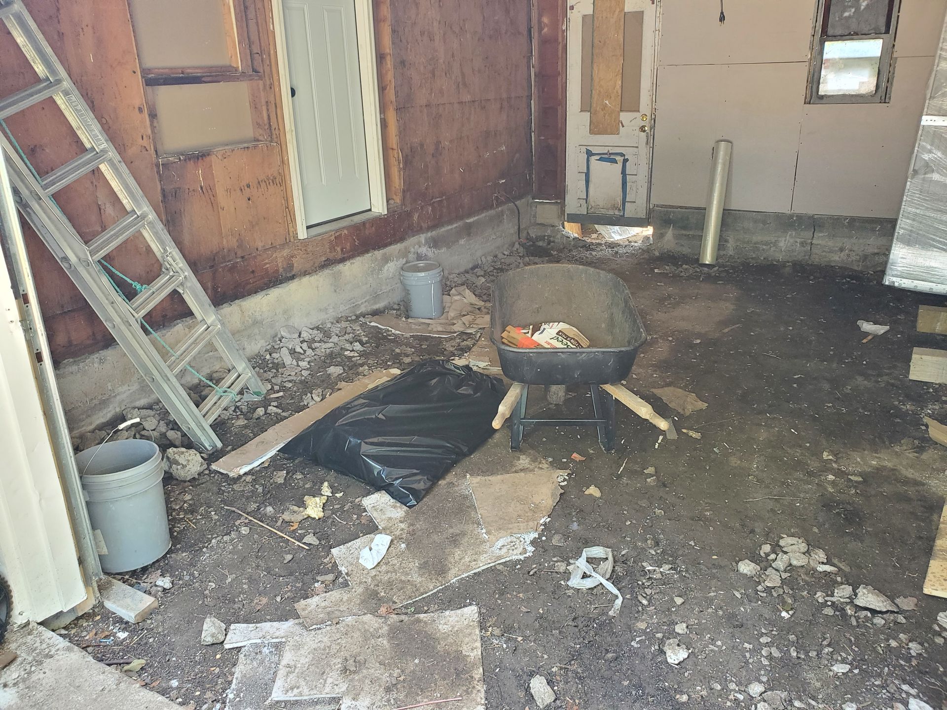 Interior view of a cluttered, unfinished room with a ladder, debris, and a wheelbarrow.