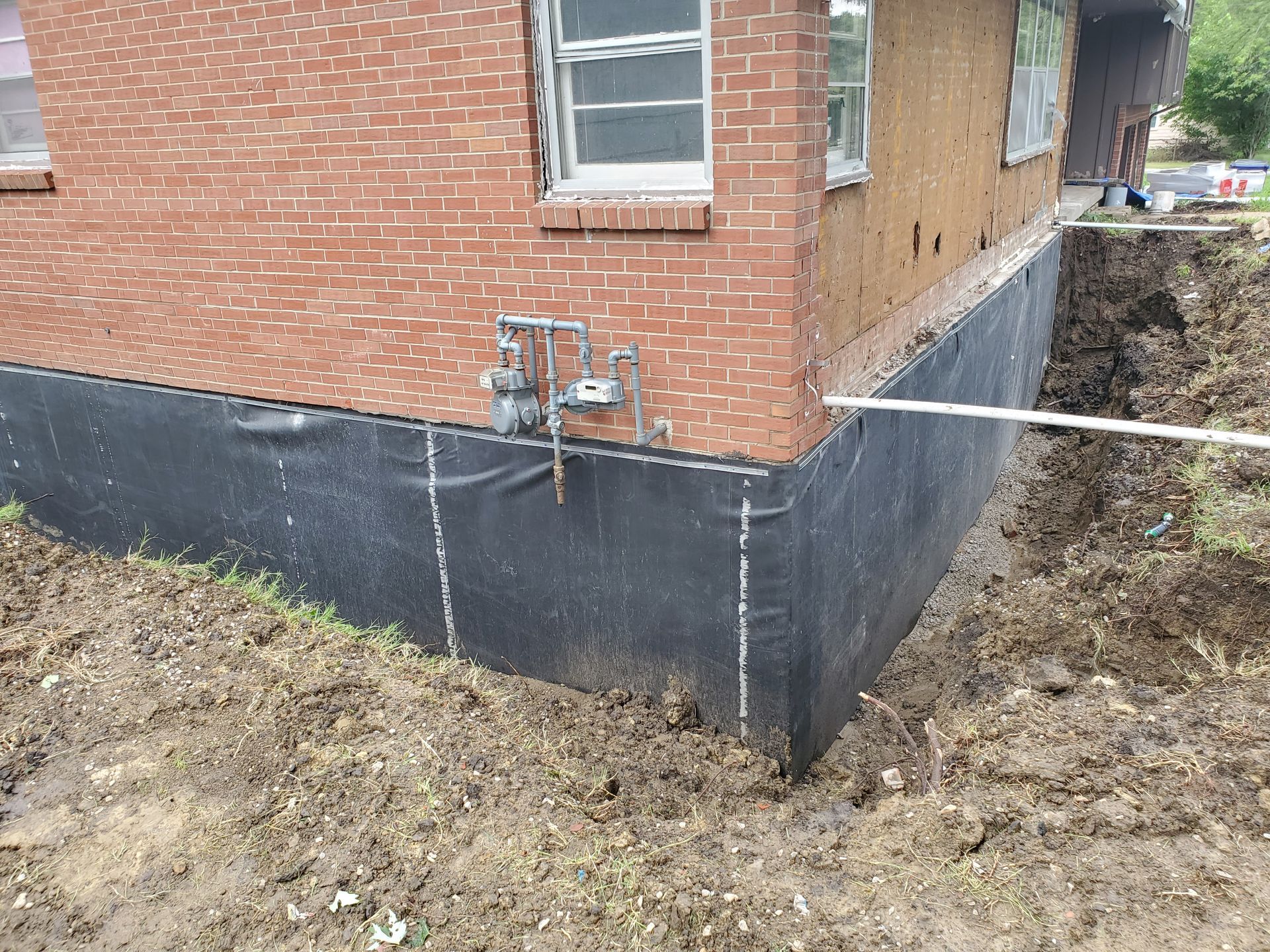 Exterior of a brick building with black protective membrane around the foundation, next to a dirt excavation.