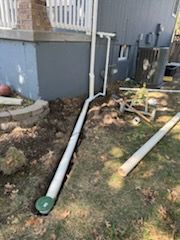 White drain pipes being installed along the side of a house on grass, soil, and a retaining wall.