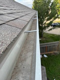 A close-up of a dirty gutter with debris. The white gutter is attached to a brown shingle roof.