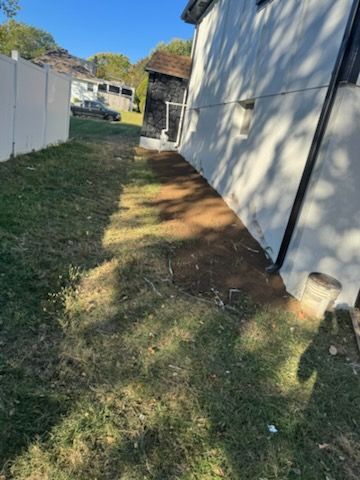 Grass and dirt area between a white building and fence. Sunny day.