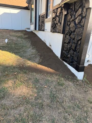 A sloped front yard with a freshly tilled strip of dirt next to a house with black stone siding.