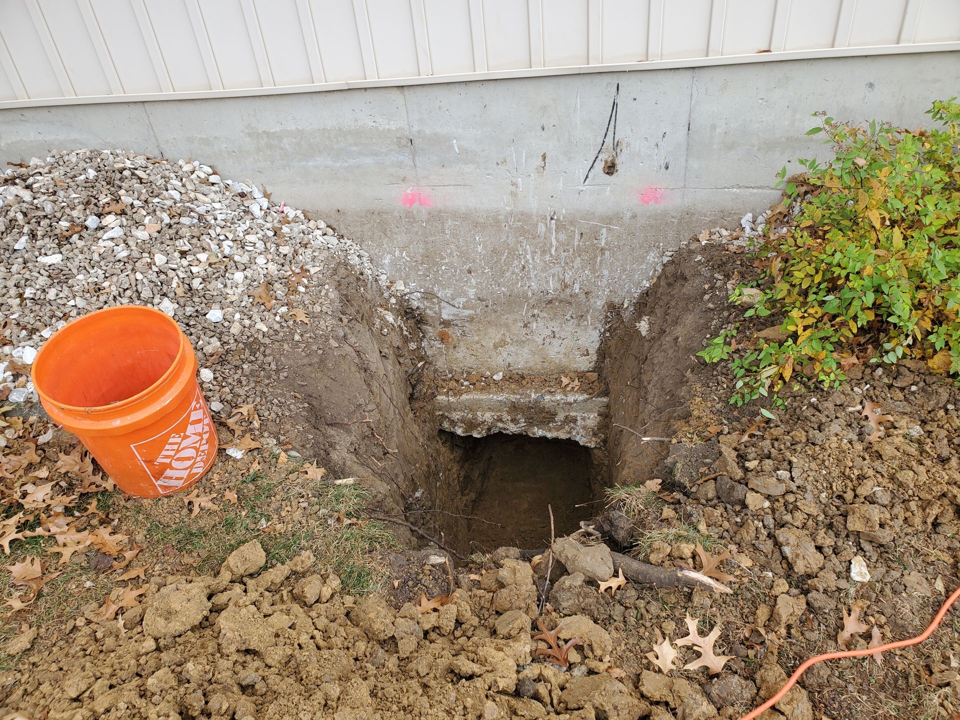 Hole dug near a house foundation; dirt, gravel, an orange bucket, and a bush are present.