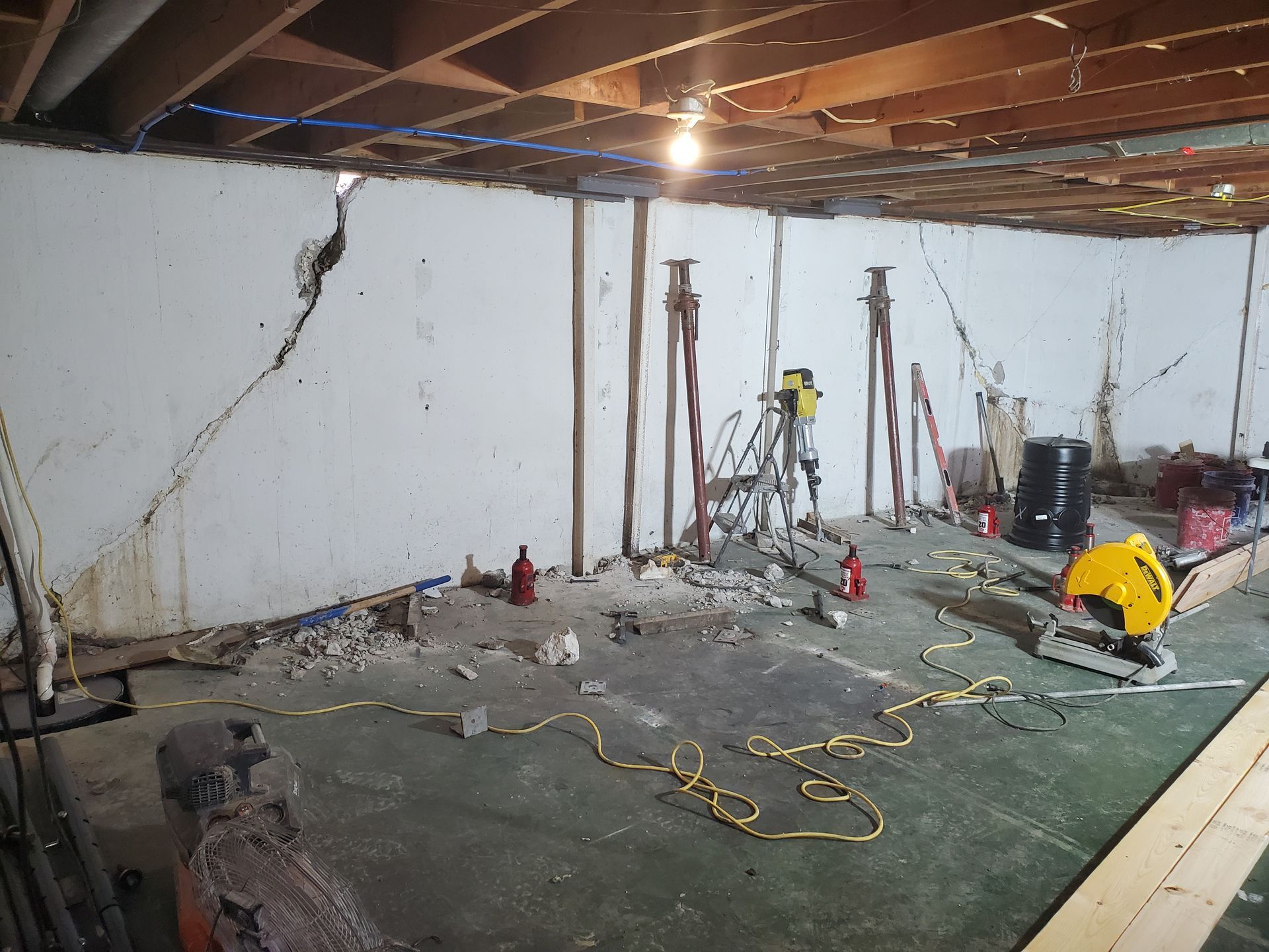 Basement with cracked white wall and construction tools, including support jacks, a saw, and a drill, in progress.