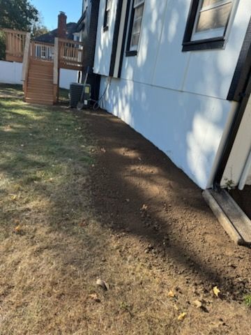 Brown soil bed along the white wall of a house; a wooden deck and green grass in the yard.