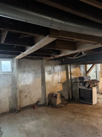 Basement interior with exposed wooden beams, ductwork, and concrete walls and floor. A window and old appliances are visible.