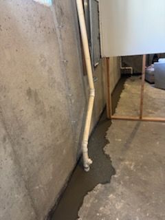 Basement with concrete wall and floor, a pipe, and a wet, gray cement-like material along the wall.