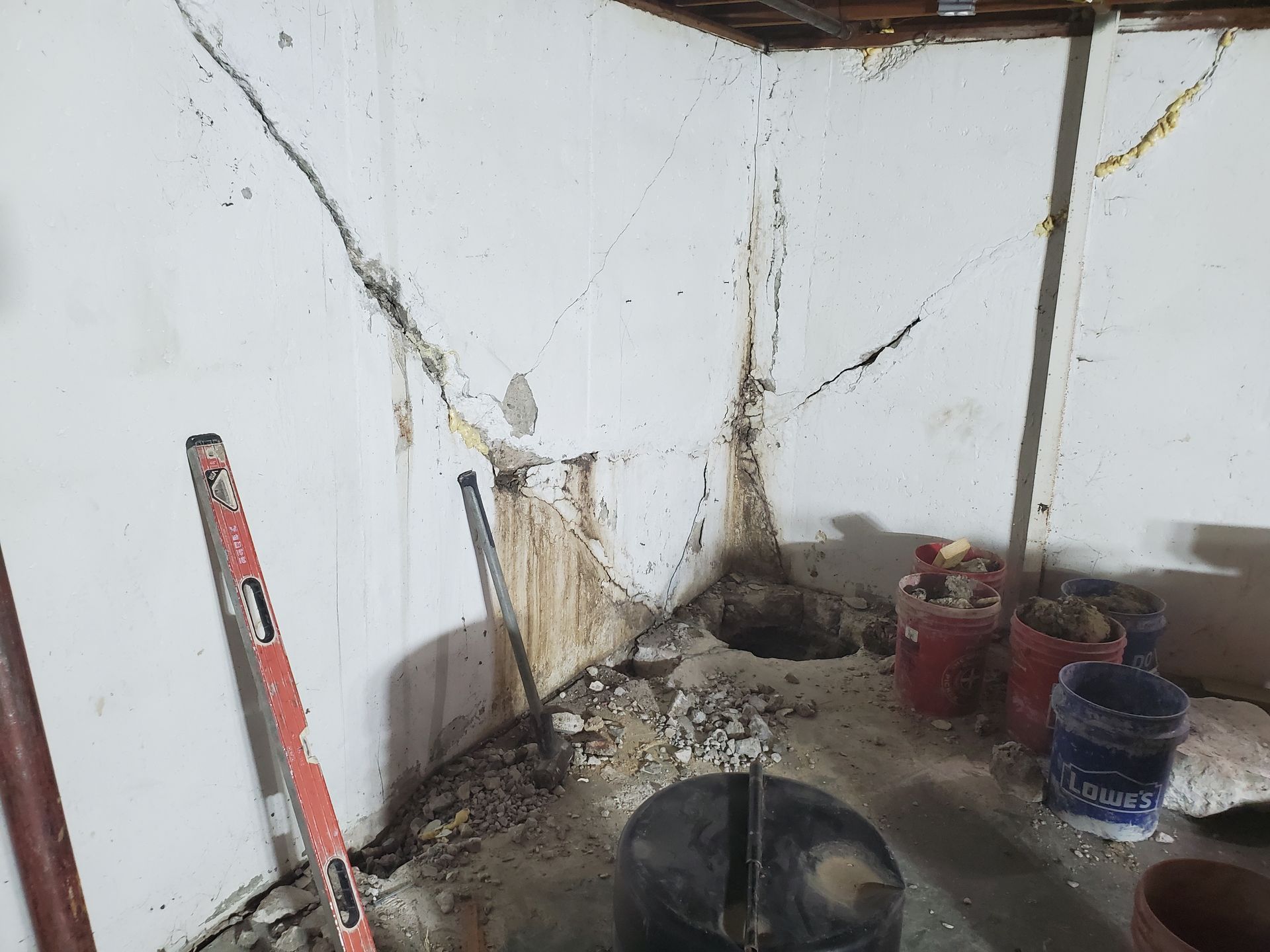 Basement wall with cracks and water damage, concrete floor, buckets, and a level.