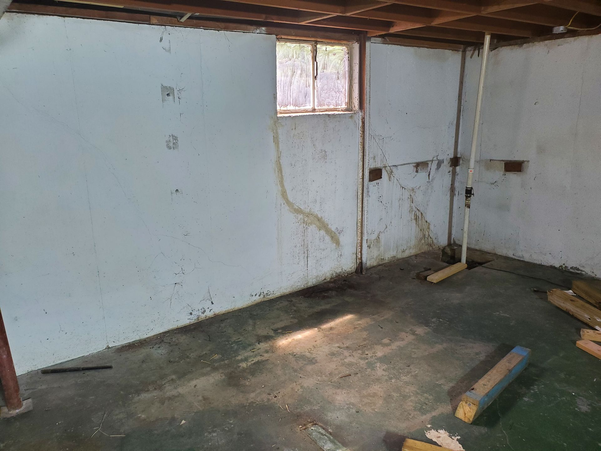 Basement interior, concrete floor, white walls with water stains, small window, wood beams overhead.