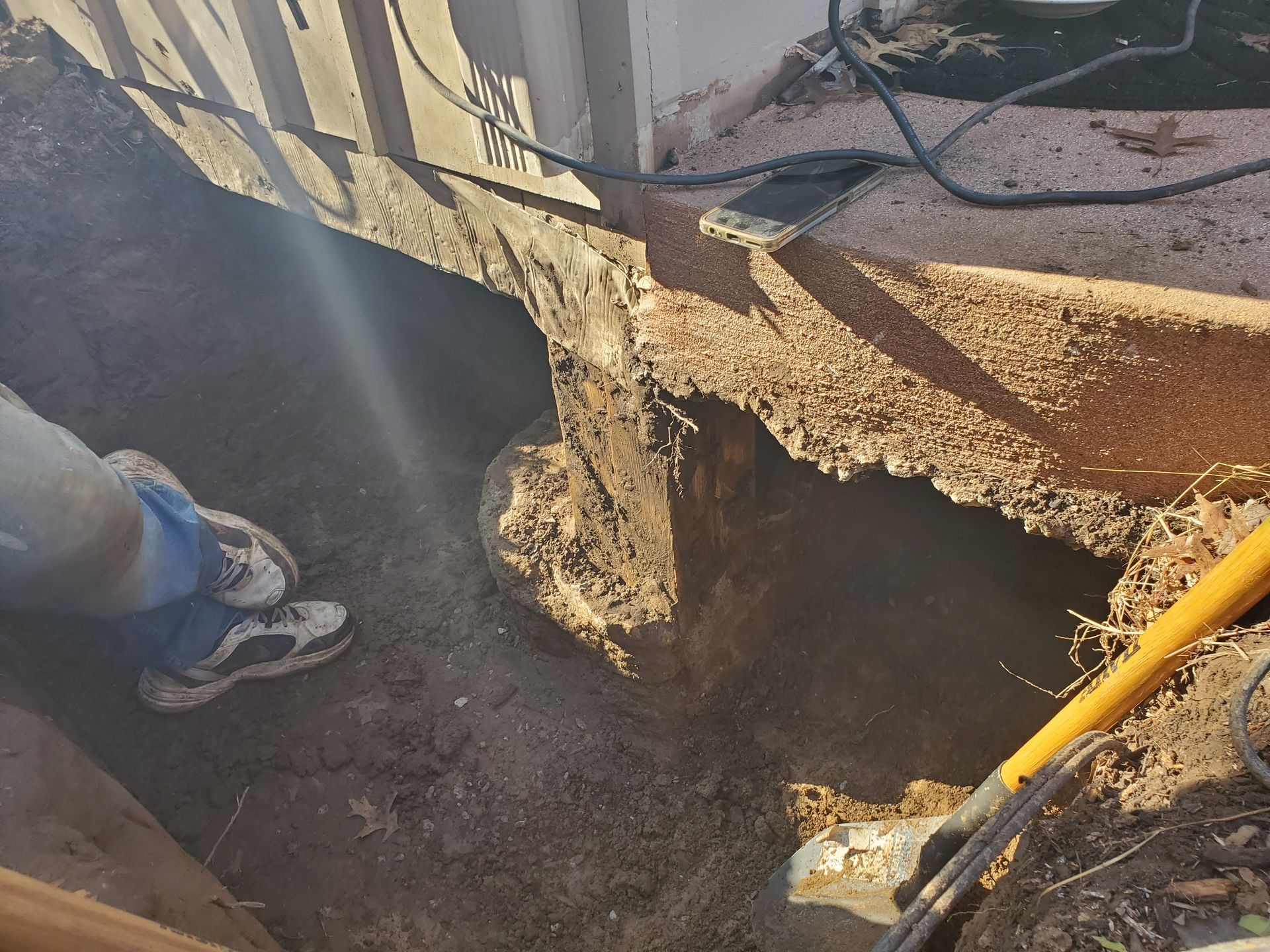 Hole dug near a building foundation; tools nearby. A person is visible from the legs down, wearing sneakers.