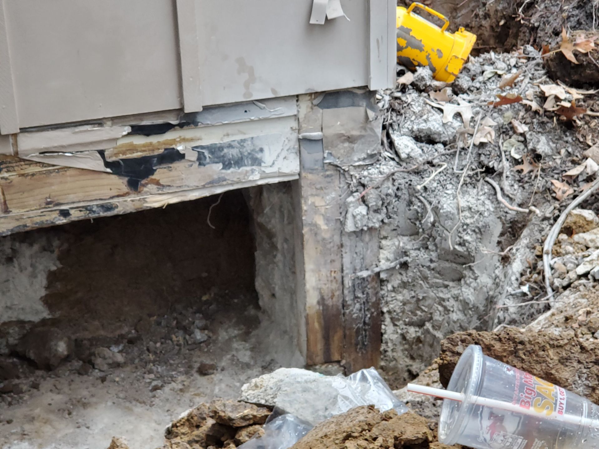Excavation exposing foundation damage near a building; soil, debris, and a yellow container are present.