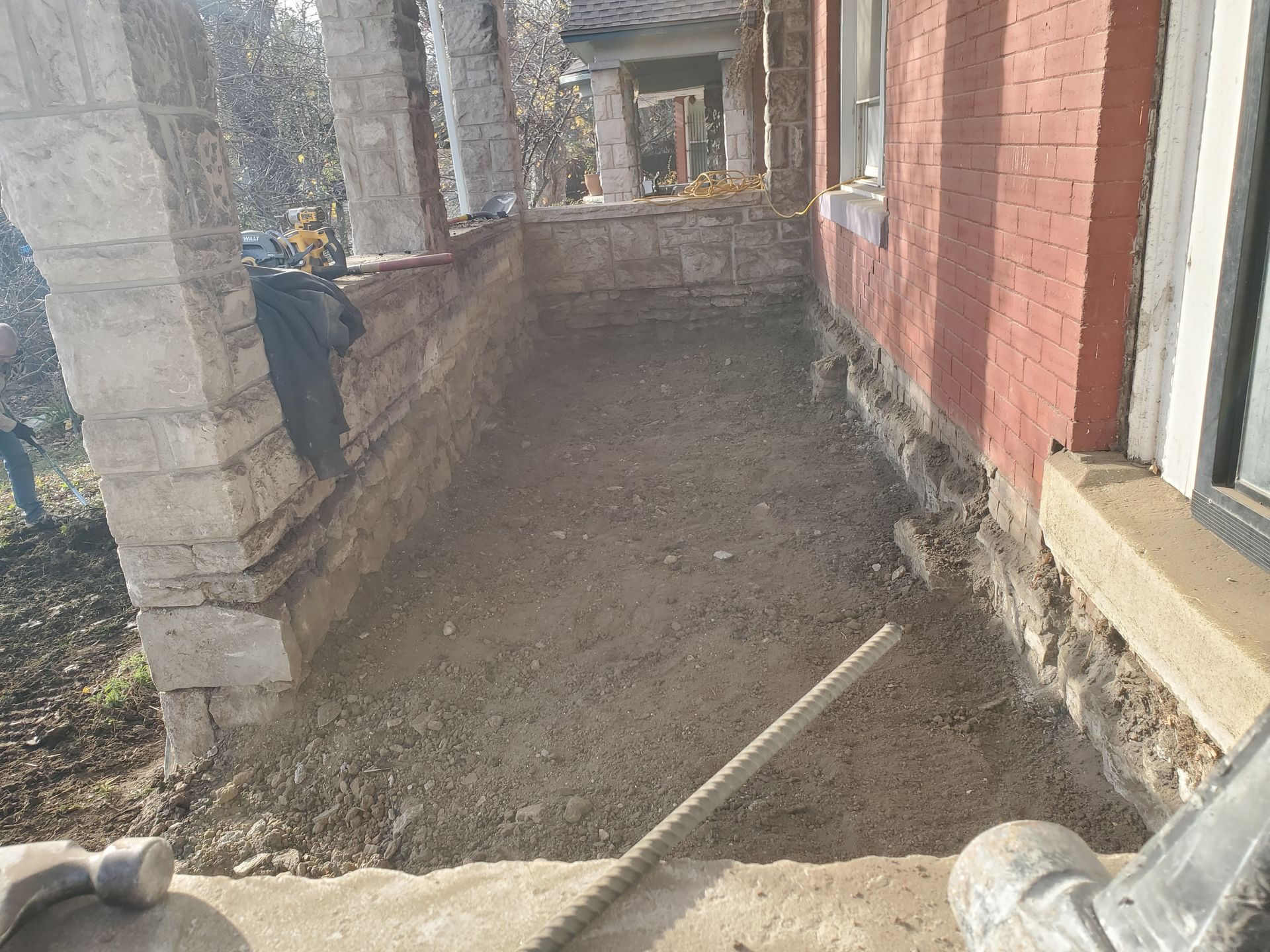 A front porch with brick and stone structures, currently dirt-filled; a long handle tool lays in the foreground.