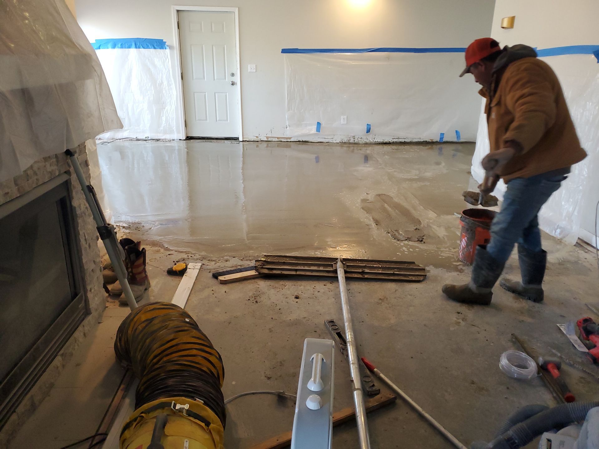 Man smoothing wet concrete floor with tools in a room, walls covered in plastic.