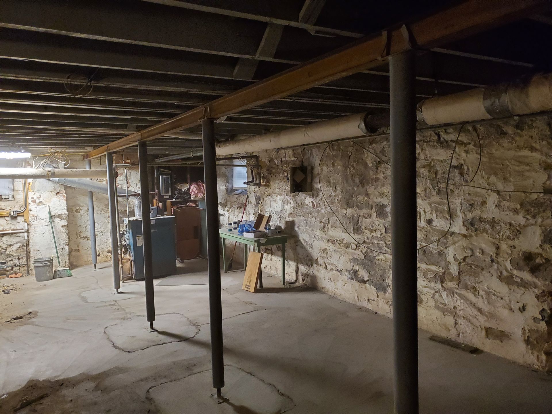 Basement with support beams, stone walls, and pipes. The floor has circular markings and is uneven.