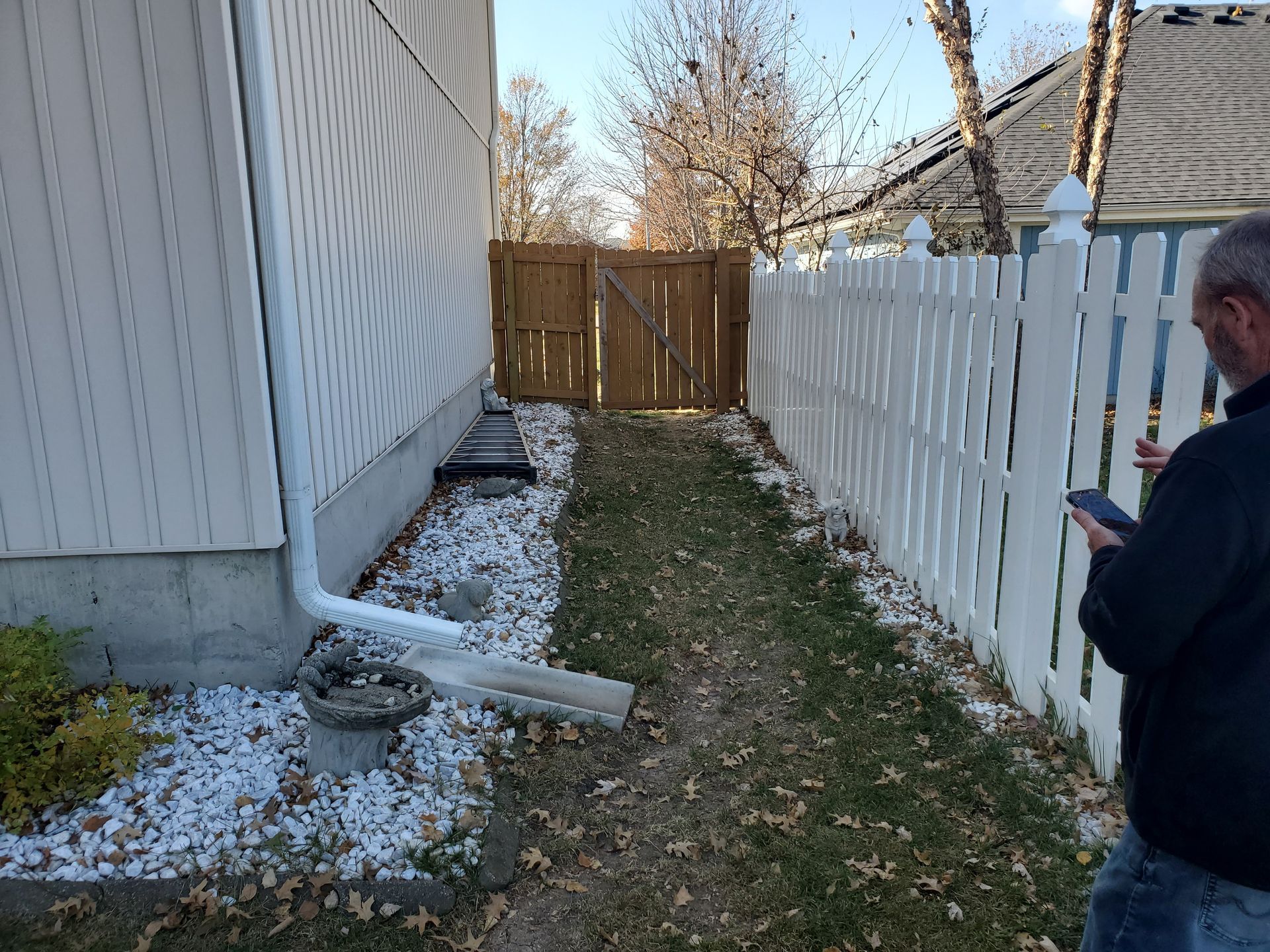Narrow grassy path between a white picket fence and a house, leading to a wooden gate; a person uses a phone.