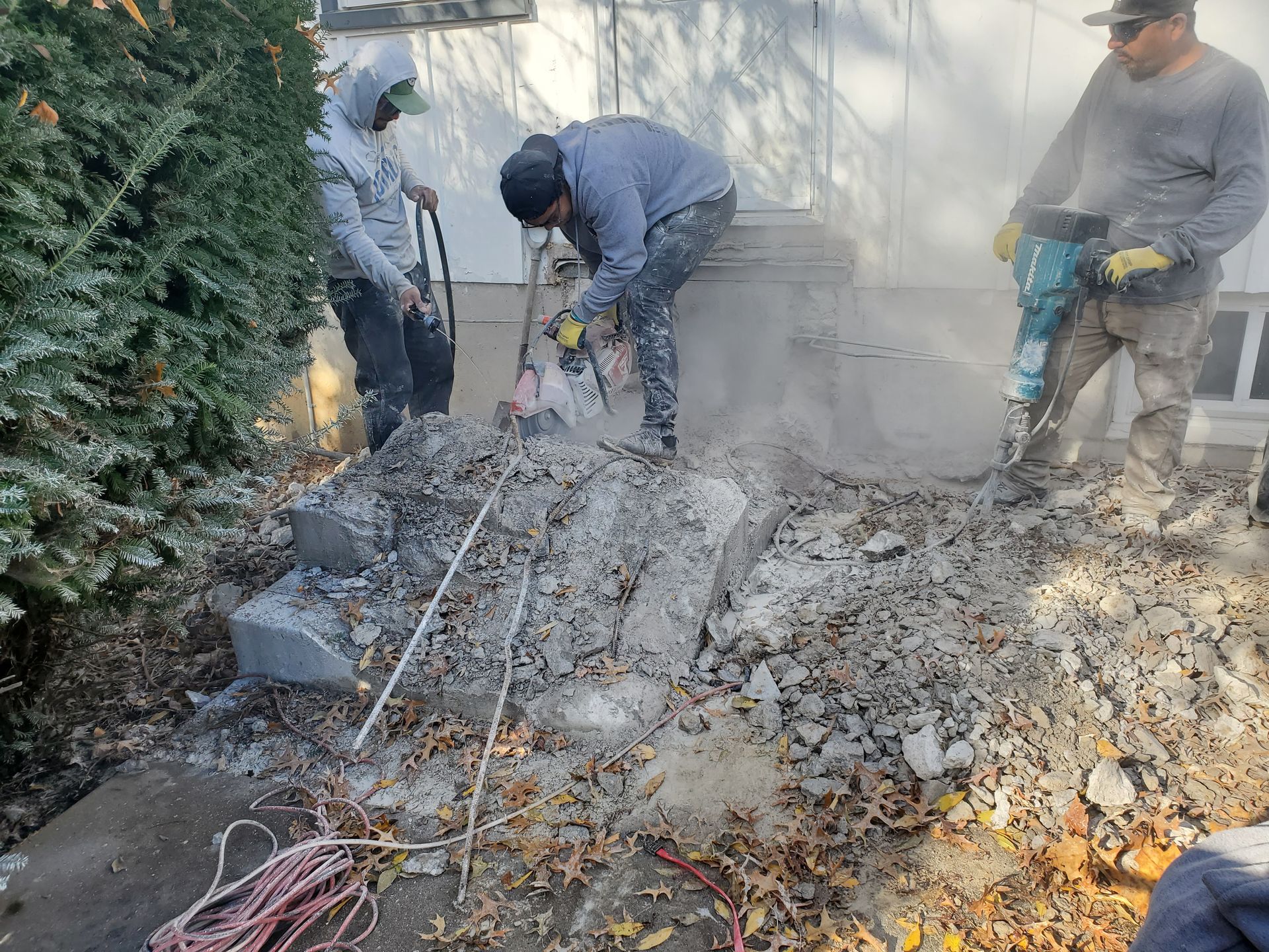 Workers demolishing concrete steps with jackhammers, creating dust. Gray concrete and debris visible.
