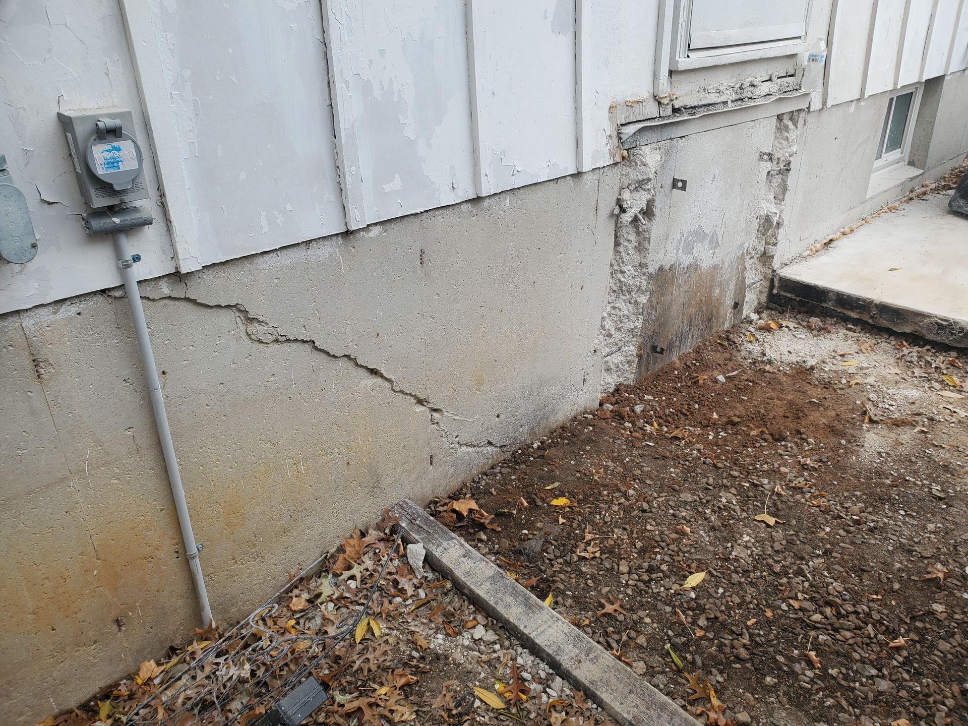 Cracked concrete foundation of a house with visible electrical meter, dirt, and gravel.