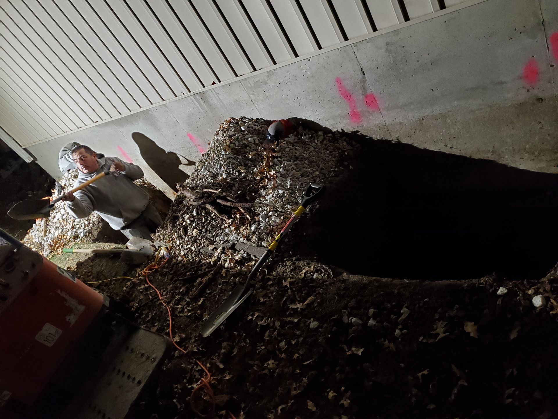 Person raking debris in a dark underpass. Concrete ceiling overhead. Orange bucket nearby. Pink markings on wall.