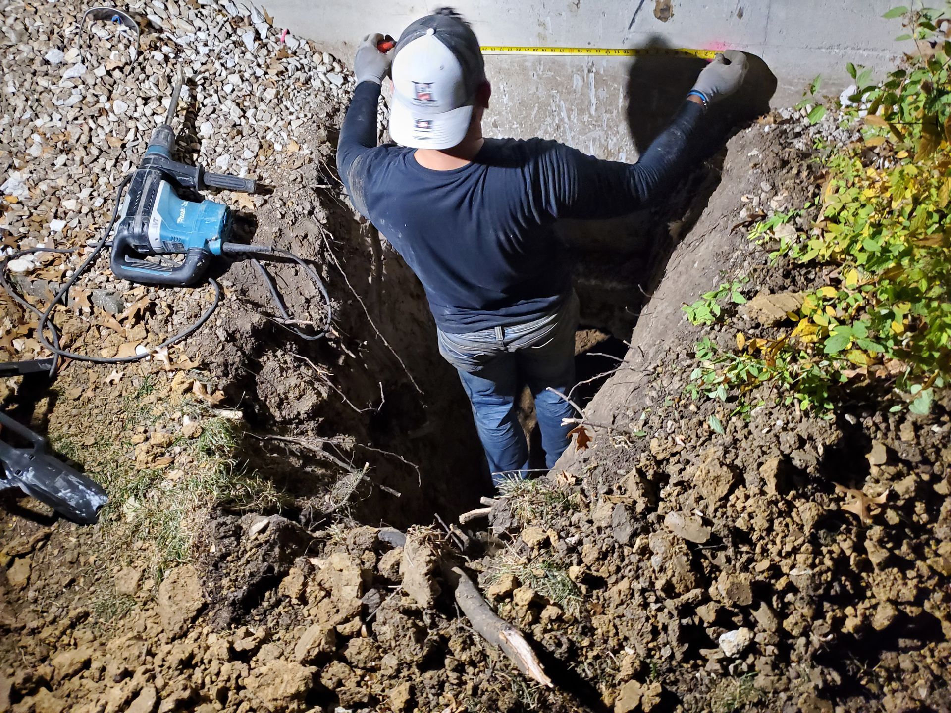 Person in a deep trench measuring, with jackhammer nearby. Dark setting.