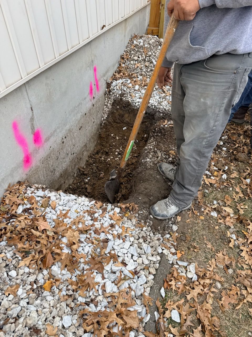 Person digging near a building foundation with a shovel. Dirt and rocks are present. Pink markings on wall.