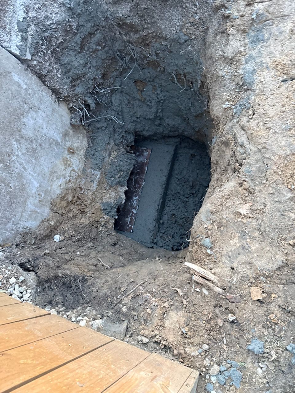 Excavation reveals a concrete rectangular structure, partially buried, next to wooden planks. Soil surrounds the opening.
