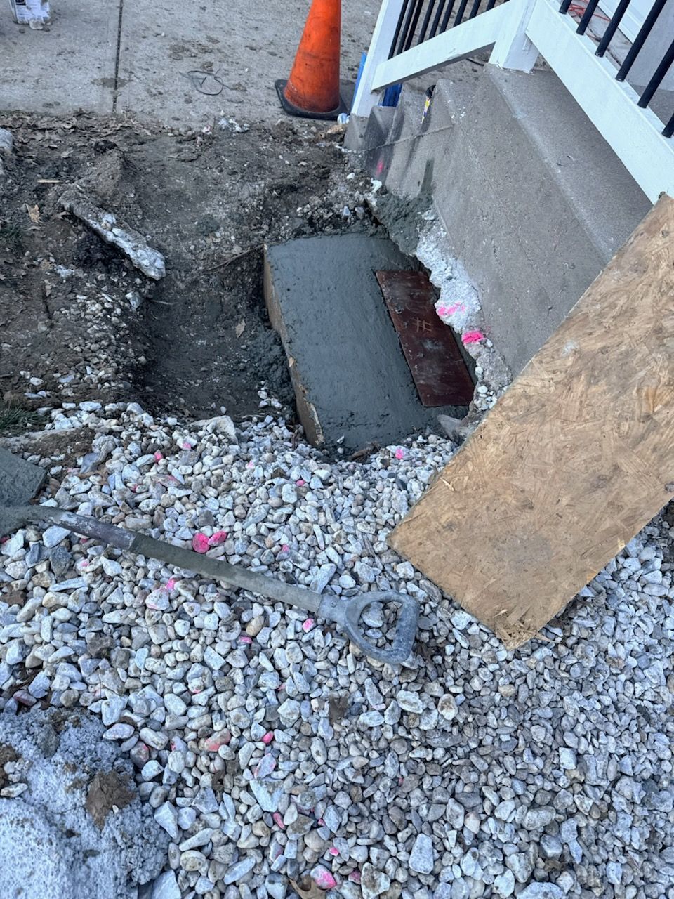 Construction site with gravel, concrete, and damaged steps; orange cone and wooden board present.