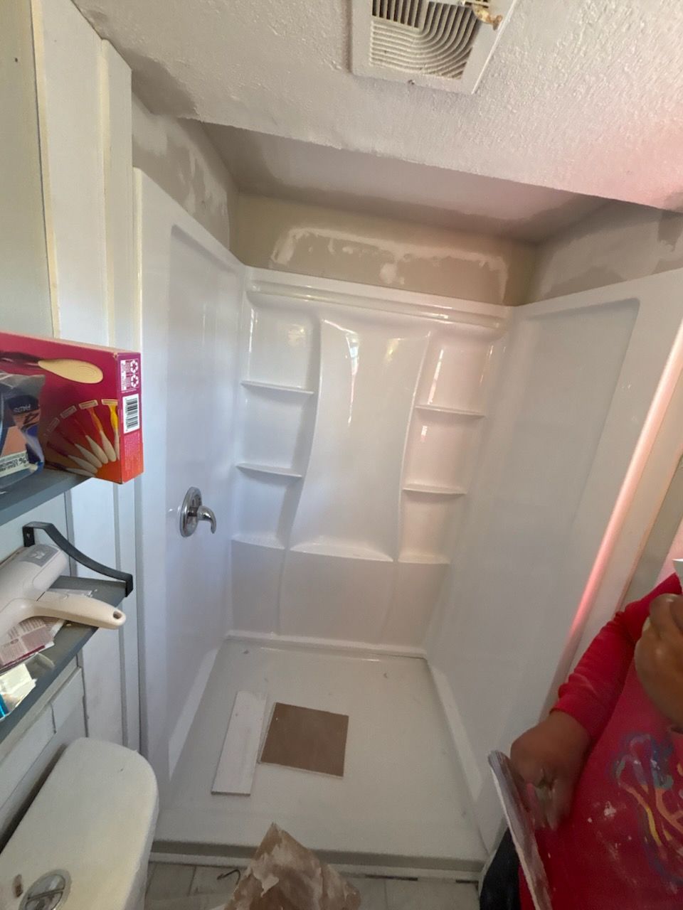 A white shower stall in a room with a partially finished ceiling. A person is visible on the right.
