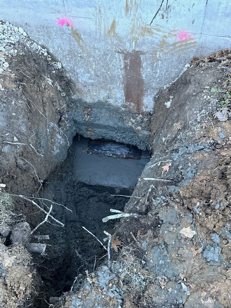 Hole dug in the ground in front of a concrete structure. Mud and debris fill the excavation.