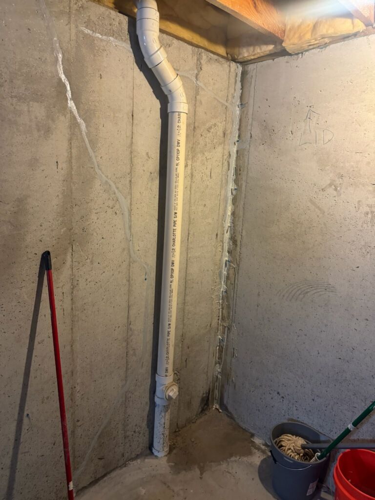 A basement interior with a tall, vertical pipe against a concrete wall. A bucket and a mop are in the corner.