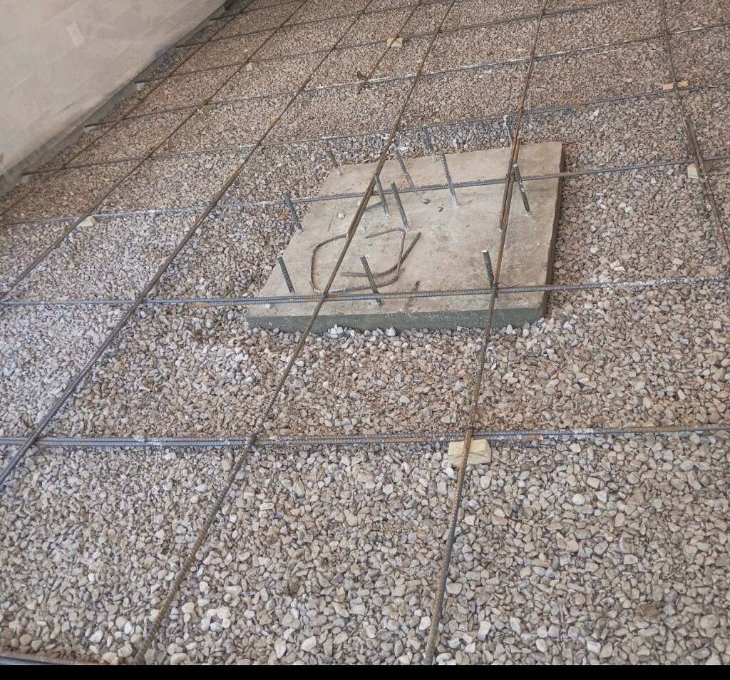 Gravel base with a grid of rebar, preparing for concrete. A square concrete block sits in the center.