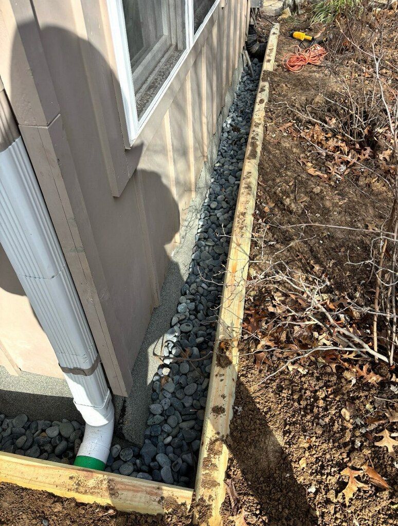 Gravel-filled trench beside a building with a white downspout.  Wooden frame borders the trench.