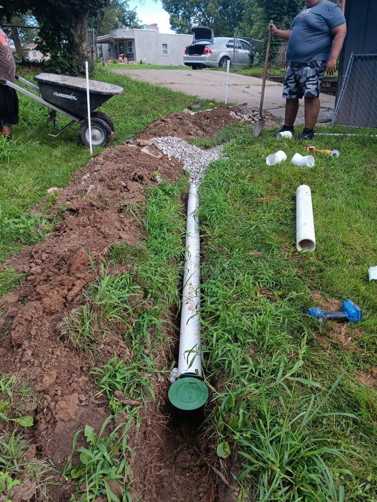 Trench dug with buried pipe and gravel for drainage on a lawn; people work in background.