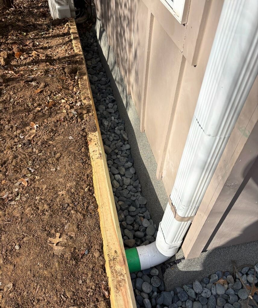 A trench filled with gravel alongside a building with a white downspout; wood planking defines the trench's edge.