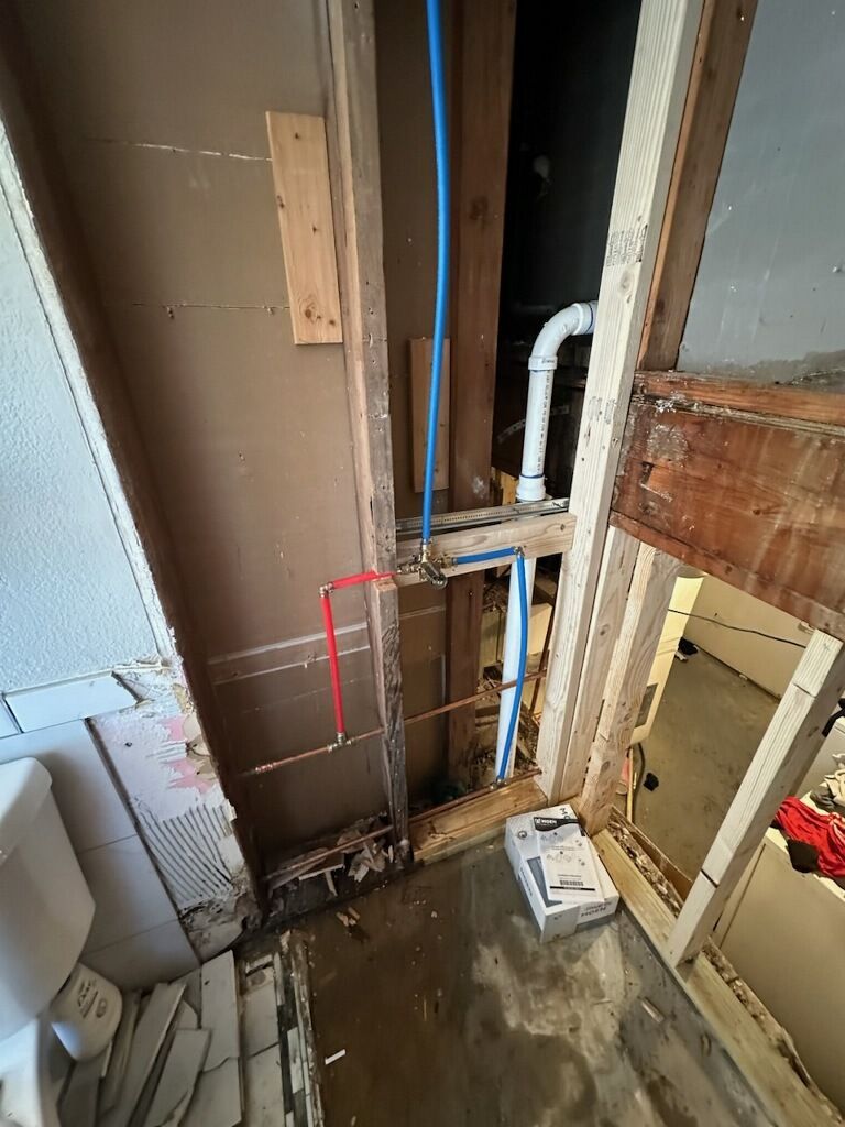 Bathroom plumbing under construction: blue and copper pipes, exposed framing, a partially demolished wall.