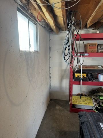 Basement corner with a window, wires, shelving unit, and tool case. Concrete walls and ceiling beams.