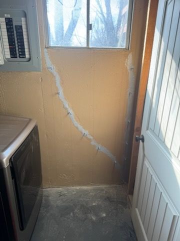 Laundry room interior with a cracked wall, window, and washing machine. A white door is on the right.